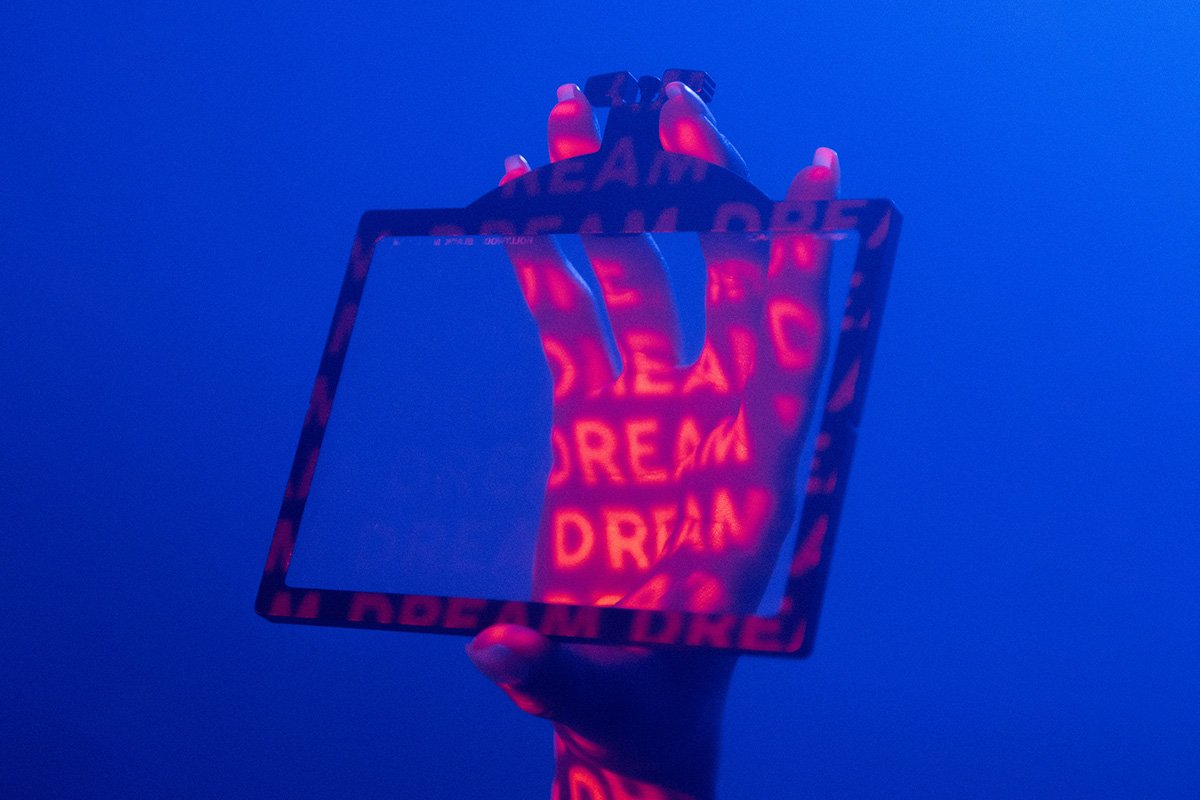 A woman’s hand in a dimly lit room, holding up a frame. The word “Dream” is projected onto her hand repeatedly.