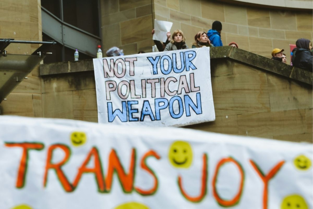 Protesters gather outside a public building during a transgender rights demonstration, holding signs and banners advocating for recognition and equality.