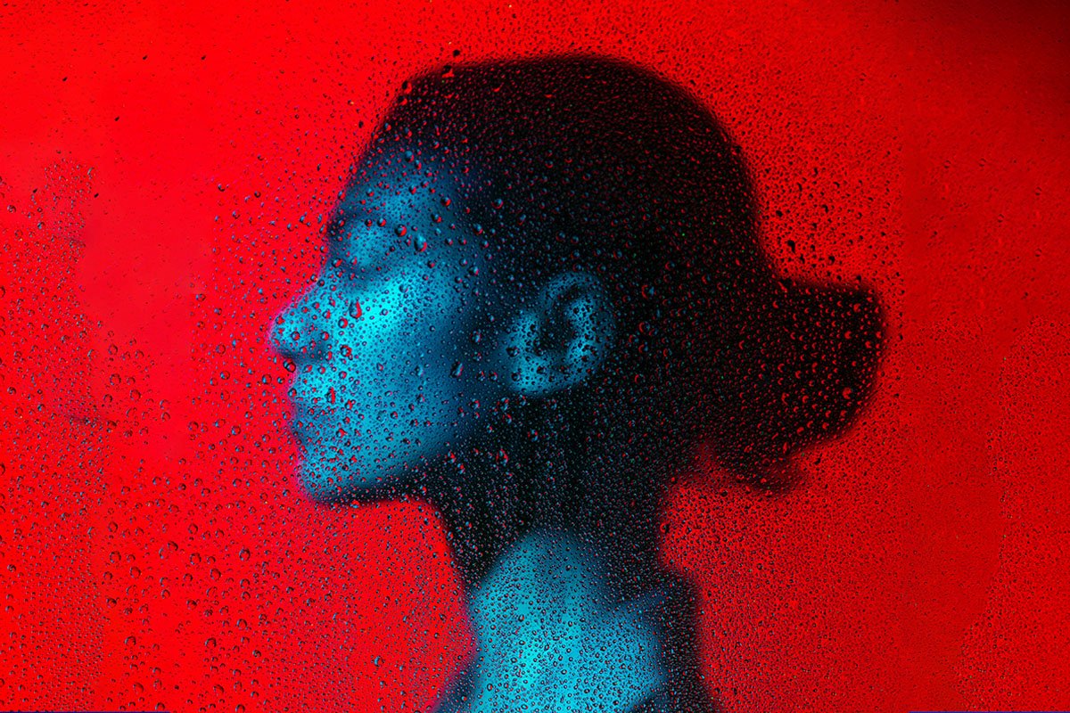 A woman with a low bun faces to the left in profile. She is washed with blue light, and the background is washed with red light. There is a film of droplets over the scene.