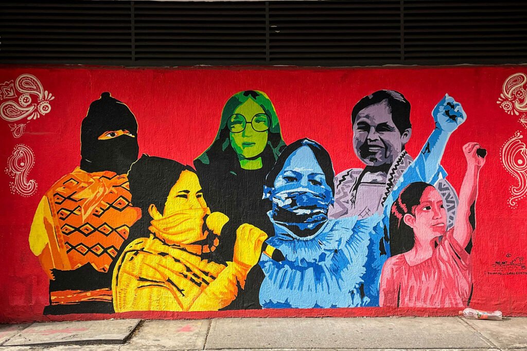 A colorful mural depicting various Mexican woman–young and elder–in various stages of activism. Two have their fists up in protest, one speaks into a microphone, one wears a balaclava, one smiles, and one gazes intensely.