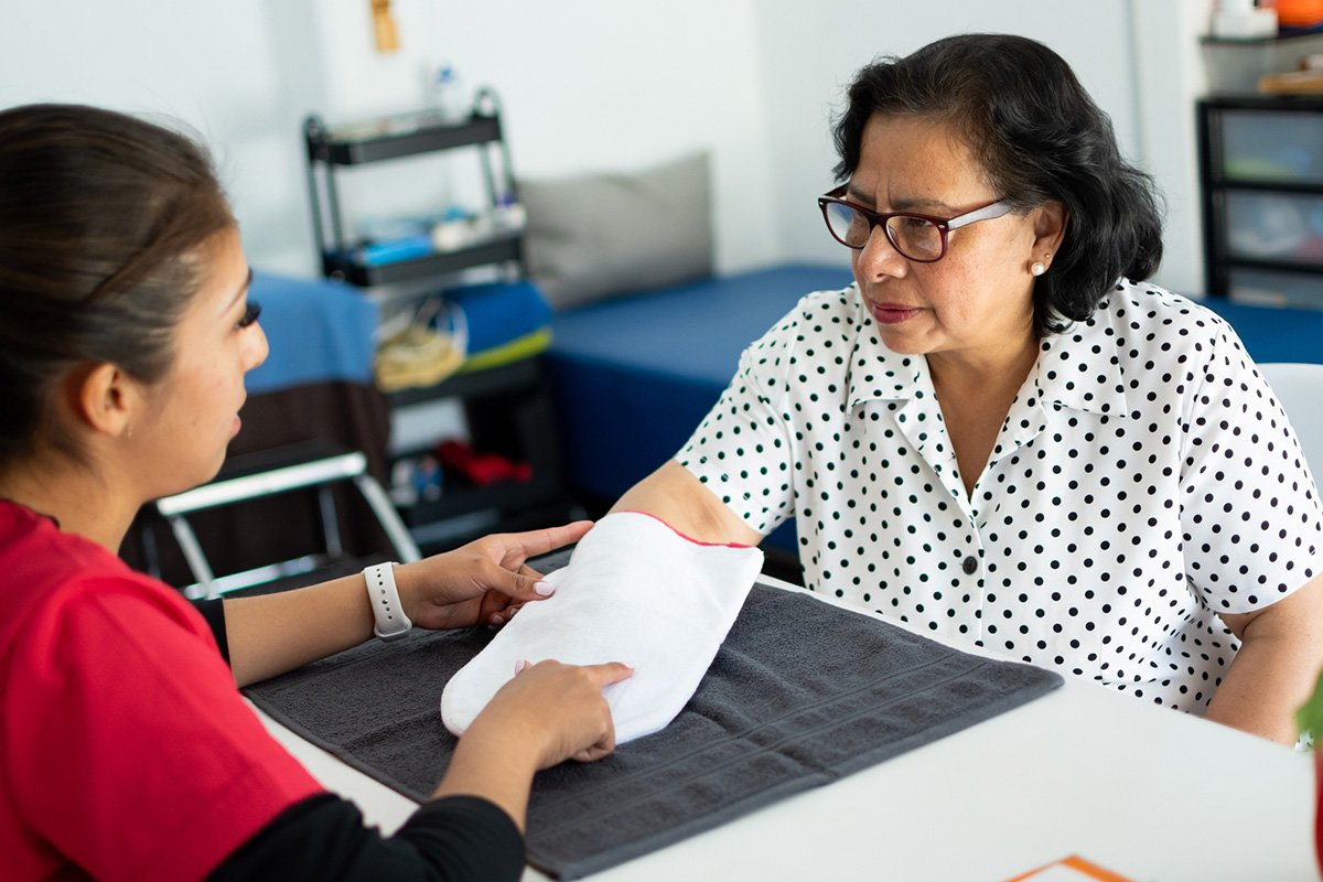 A brown-skinned elderly Latina woman with short wavy hair sits across from a medical professional who is adminsitering an arthritis therapy.