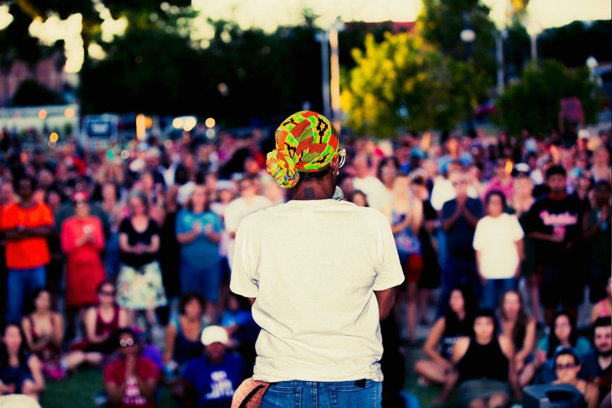 Seen from behind, a woman in a headwrap and white t-shirt stands before a diverse crowd in an outdoor gathering at dusk.