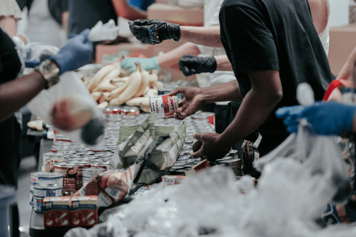 Volunteers wearing gloves sort canned goods, bananas, and packaged food at a community food distribution table.