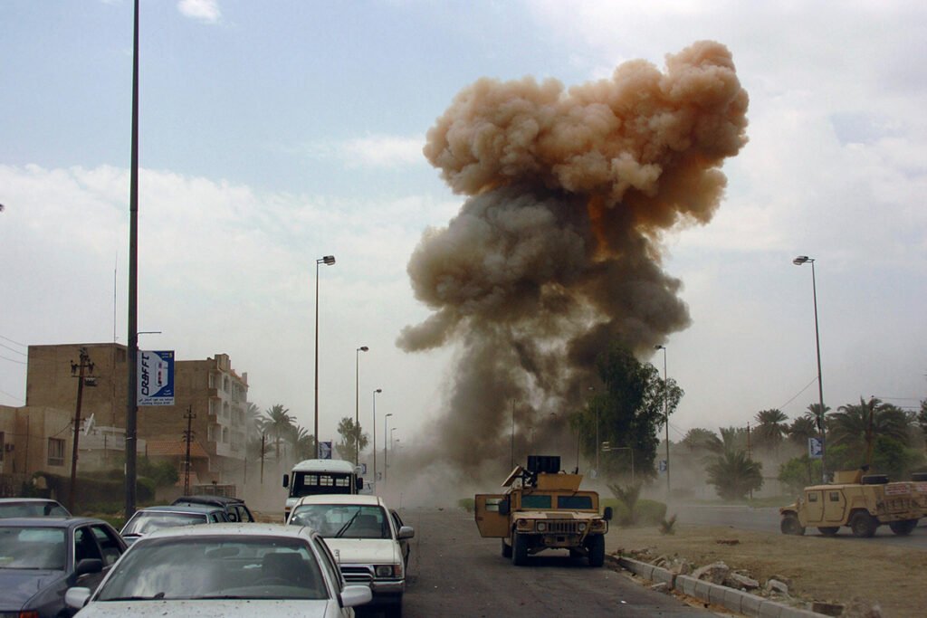 An photograph of an active car bombing scene with army tanks and civilian vehicles nearby.