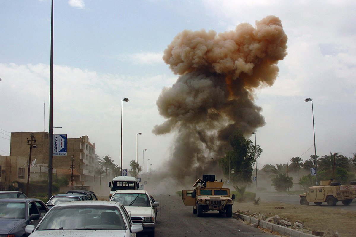 An photograph of an active car bombing scene with army tanks and civilian vehicles nearby.