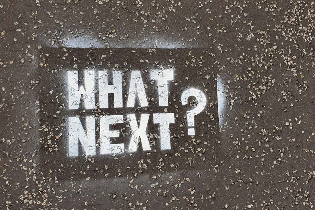 A stenciled graffiti reading, “What Next?” on a textured gray surface.