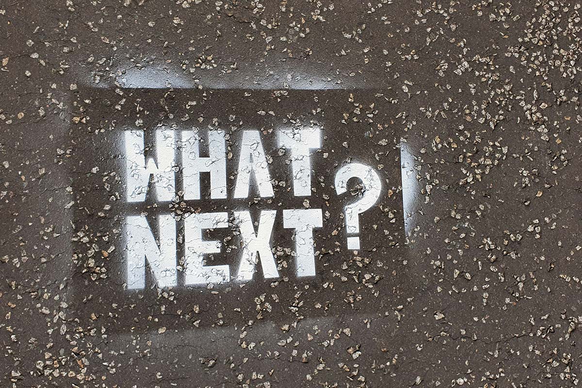 A stenciled graffiti reading, “What Next?” on a textured gray surface.