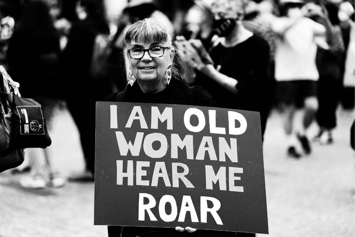 A smiling older woman in glasses holds a sign reading "I Am Old Woman Hear Me Roar" at a protest, photographed in black and white with marchers blurred behind her.