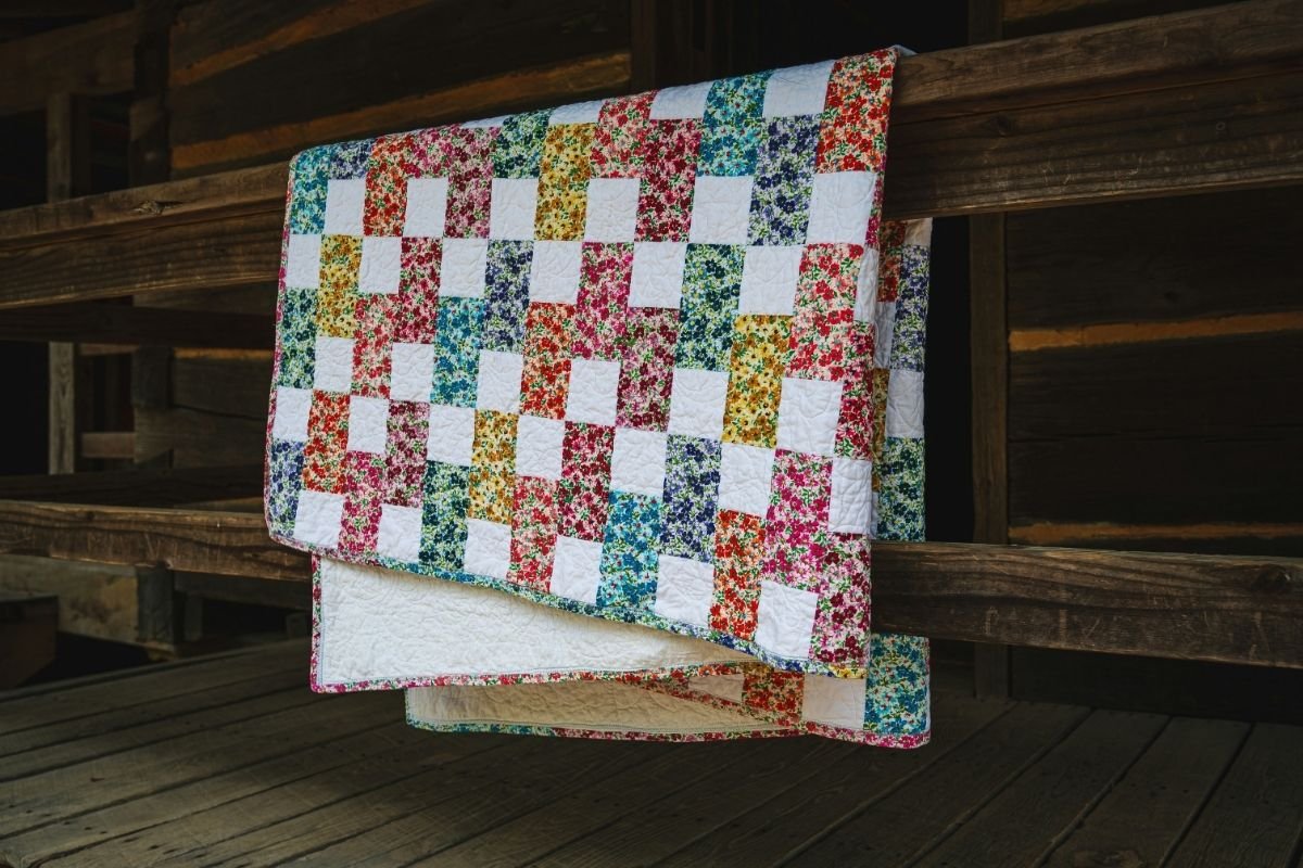 Colorful handmade patchwork quilt featuring floral fabric squares and white blocks draped over a rustic wooden railing in a barn-like setting. The quilt displays a vibrant mix of pink, blue, yellow, purple, and orange floral patterns against a dark wood background.