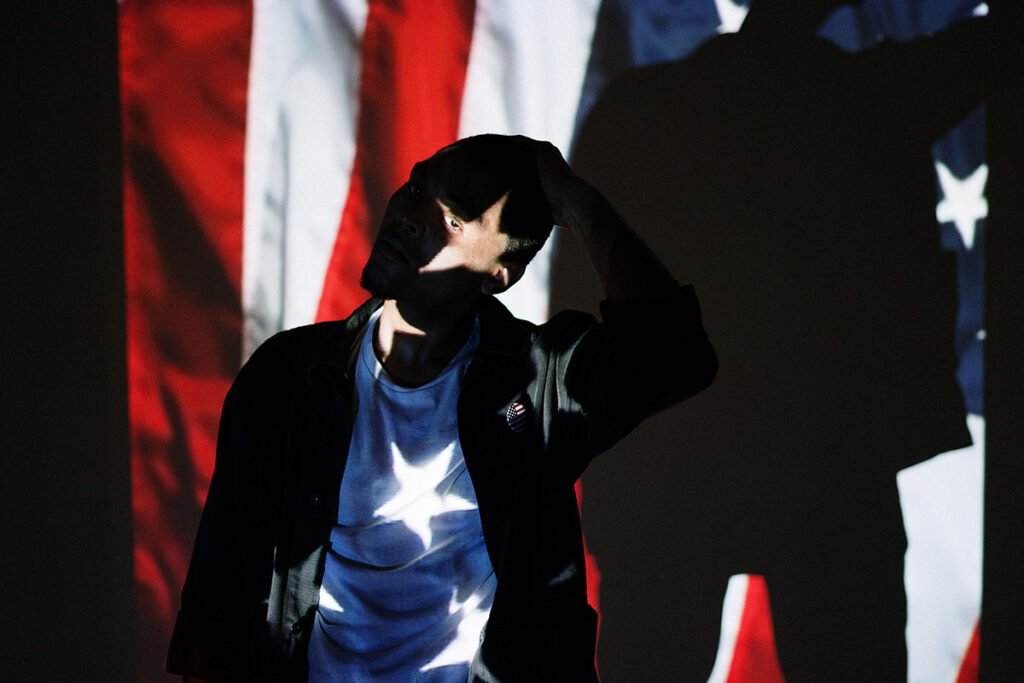 A Black man stands in a dark room where an American flag is projected over his body. He leans his head to the side with his hand atop his head. A star frames his eye as he looks at the camera.