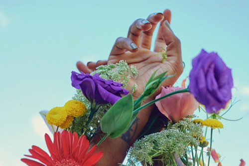 An image of purple, yellow, red flowers and greenery with a hand emerging and reaching upwards.