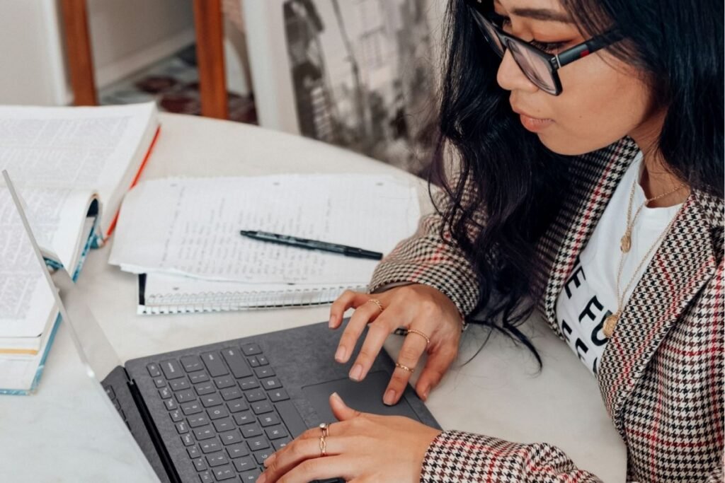 A woman in glasses and a houndstooth blazer works on a laptop at a desk surrounded by open notebooks and papers.