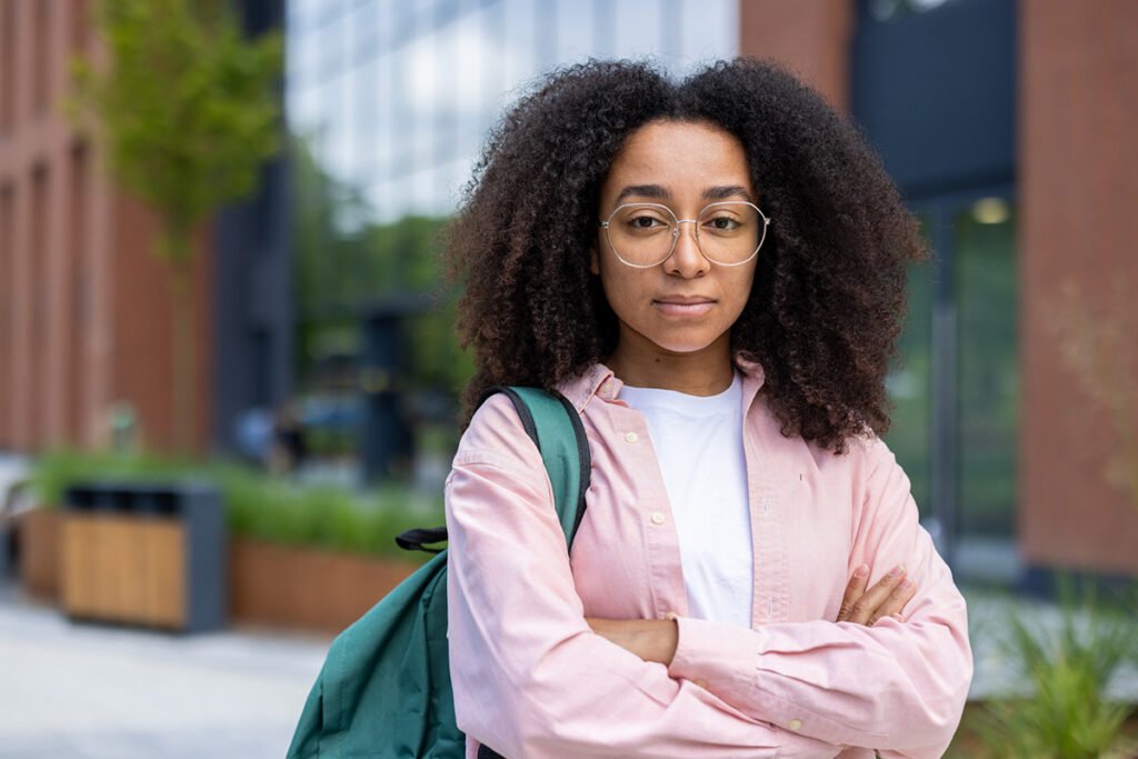 Portrait of a young female student with curly hair, and brown skin standing outdoors on a college campus holding a backpack. Her expression is serious.