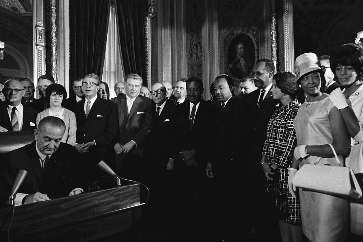 A black and white photo of President Lyndon B. Johnson, a serious-looking man with slicked hair, signs the Voting Rights Act in 1965. Behind him, several notable Civil Rights activists stand behind him, including Martin Luther King Jr.