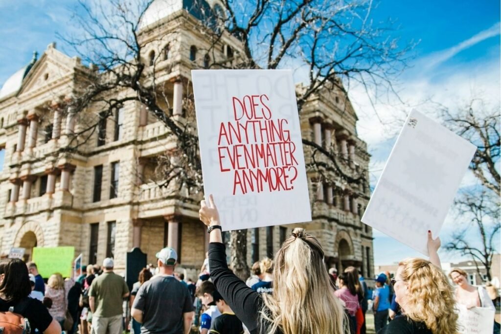 A protester holds a handwritten sign reading "Does anything even matter anymore?" above a crowd gathered outside a government building.
