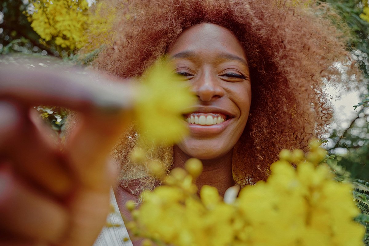 A Black woman with auburn afro-textured hair smiles broadly as she holds a small yellow flower up to the camera. There are yellow flowers in the foreground.