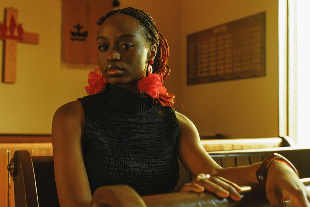 A young Black Woman with red flower earrings sits in a church pew and looks into the camera with a serious look on her face. In the background is a wooden cross.