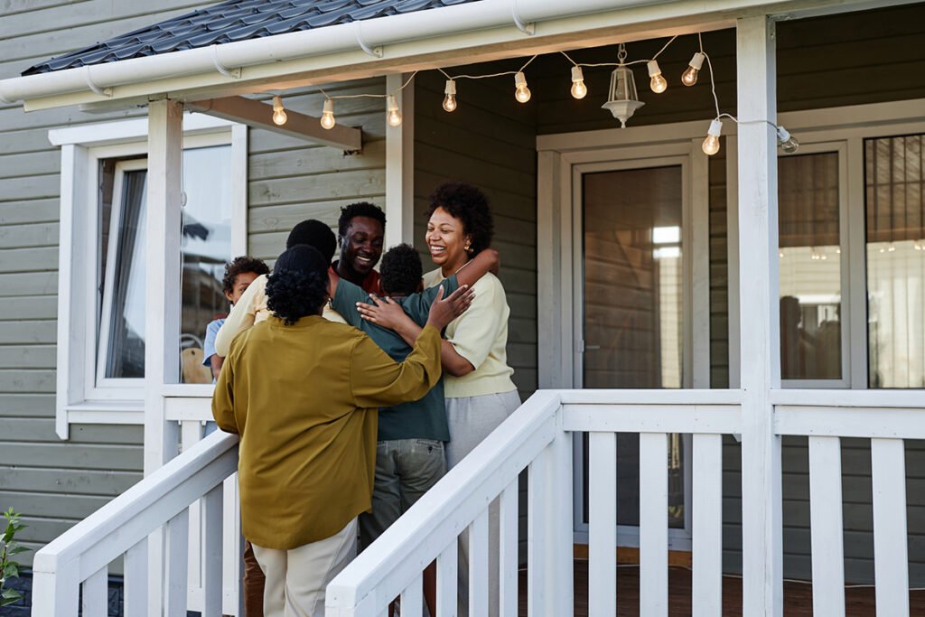 A multi-generational Black family embraces on the porch of a painted wood house. They smile and hold each other.