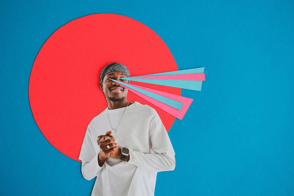 A paper collage of a smiling Black man clasping his hands and looking off into the distance. Behind him is a halo of red, and multicolored rays of paper smit from his eyes, symbolizing vision.