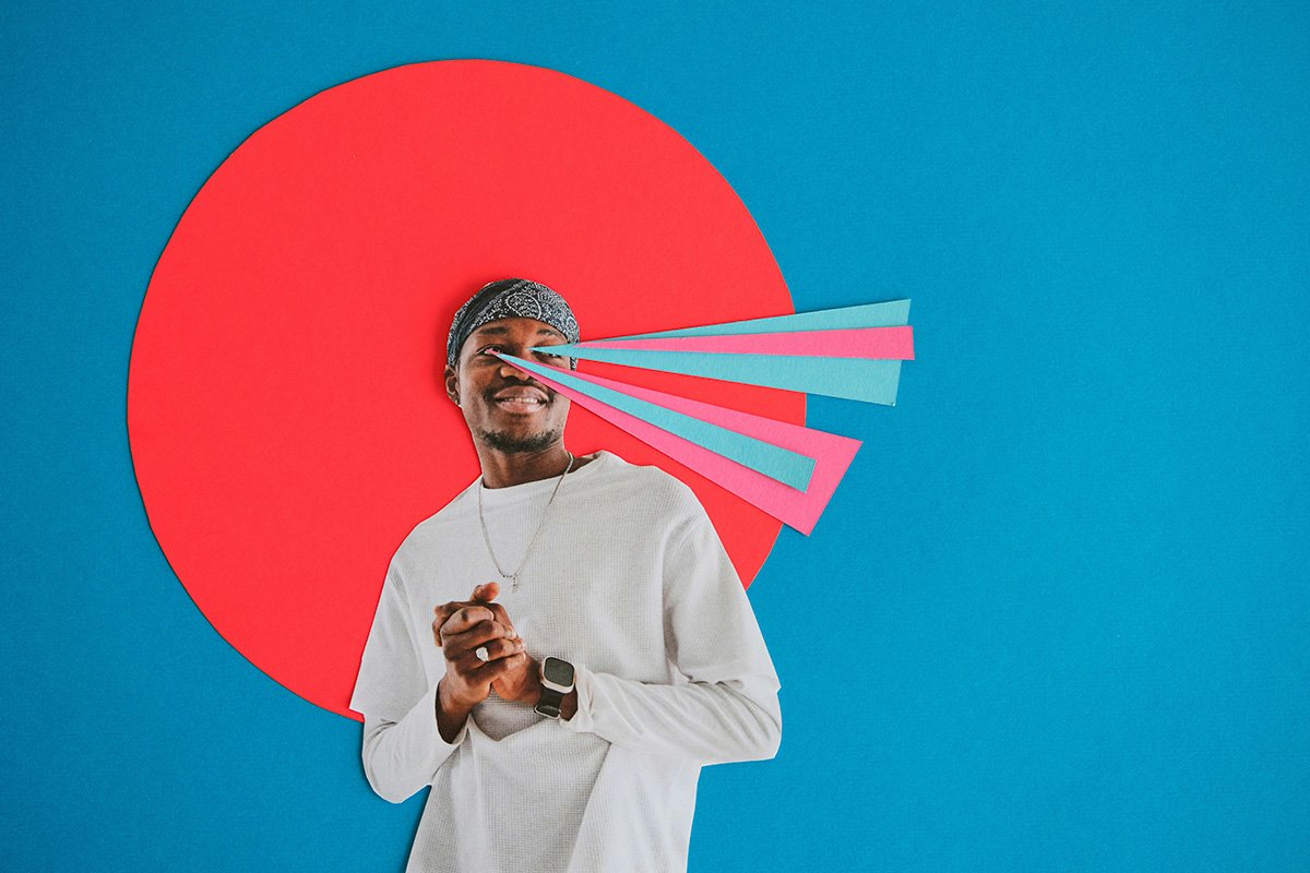 A paper collage of a smiling Black man clasping his hands and looking off into the distance. Behind him is a halo of red, and multicolored rays of paper smit from his eyes, symbolizing vision.