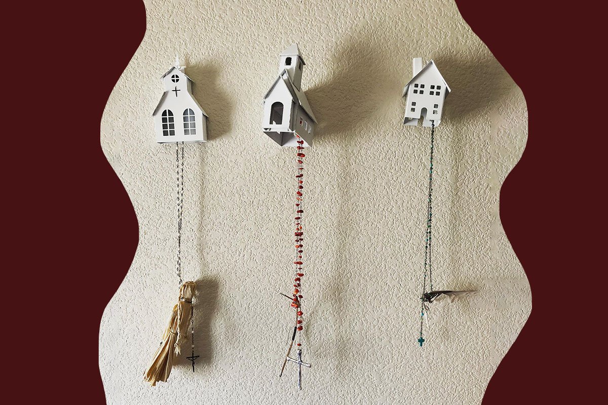 Art installation of three toy buildings, including a church, schoolhouse, and house, with rosary beads hanging from each structure. The church has a corn husk dangling from the end of the rosary, while the schoolhouse has a bow and arrow, and the house a feather.