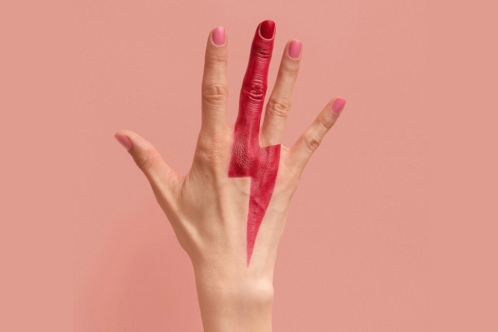 A woman’s hand with a red lightning bolt painted down the middle finger onto the back of the hand. The hand is raised against a pink background.