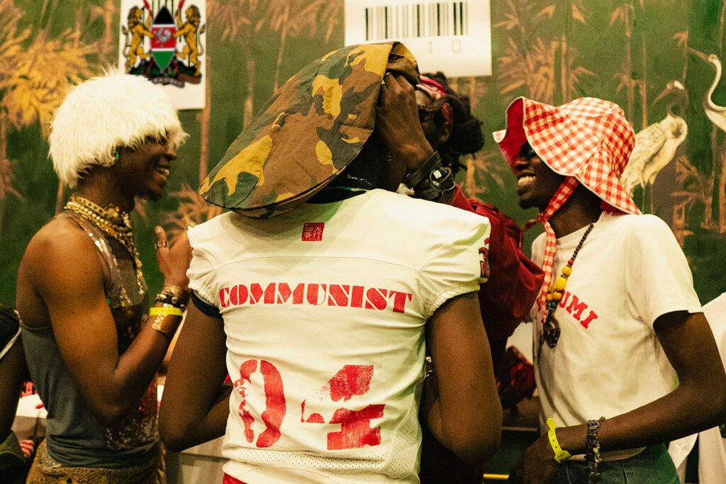 A group of fashionable youth activists in Nairobi, Kenya at a community event. One attendee wears a fur hat, another a shirt reading “communist” and another a checkered bucket hat. On the wall behind them is the Kenyan coat of arms, which features the word “Harambee” as a central part of Kenyan culture.