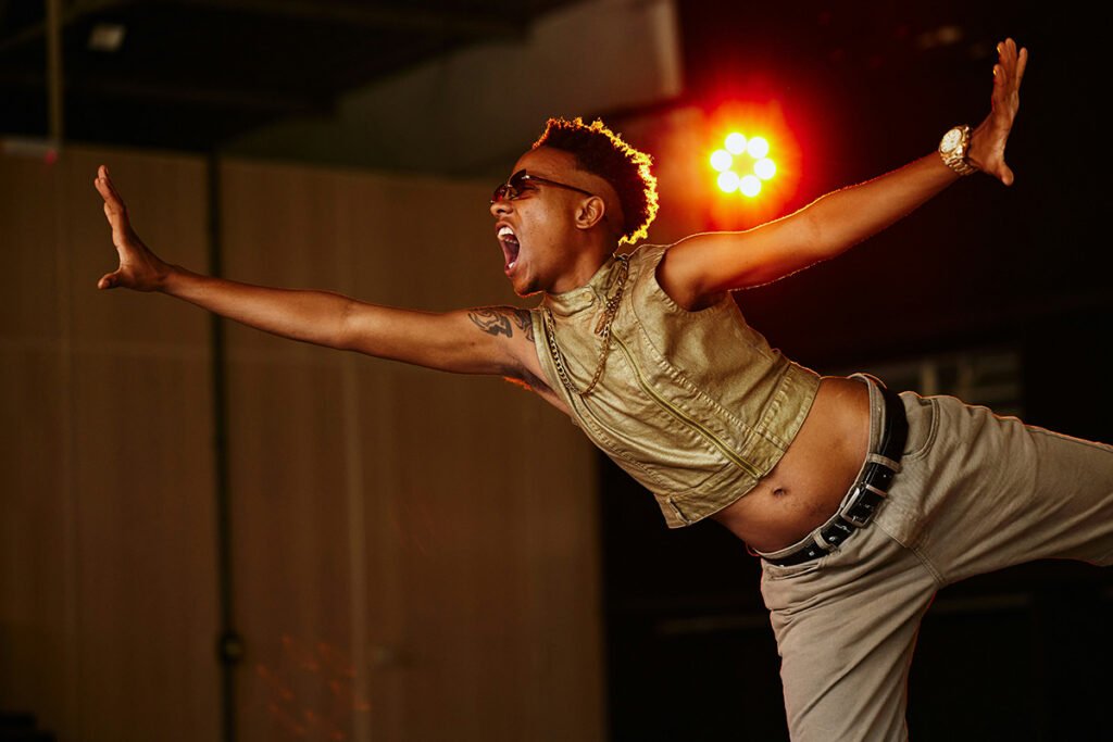 A Black person with a mohawk-afro striking an extended dance pose on a lit stage. There are rafters in the background.