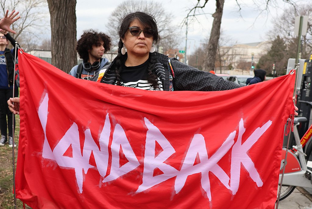 A native woman with sunglasses and two briads holds a red banner reading “Landback”
