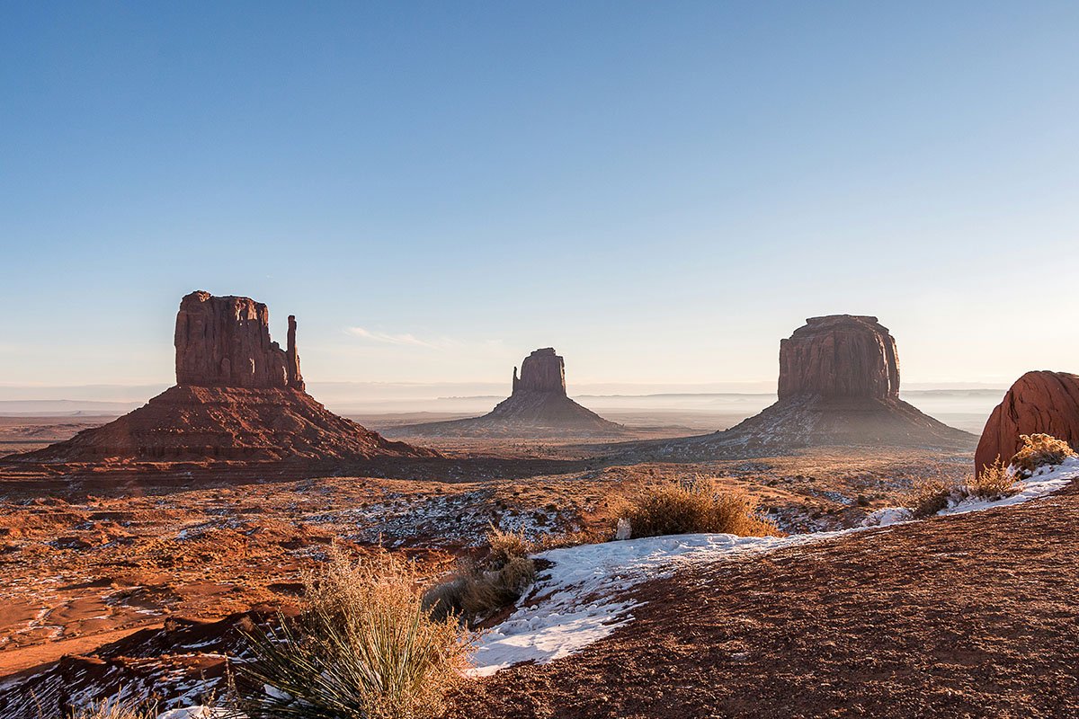 A picturesque landscape of arid western American land, dotted with tall natural structures, small bushes, and peppered with snow.