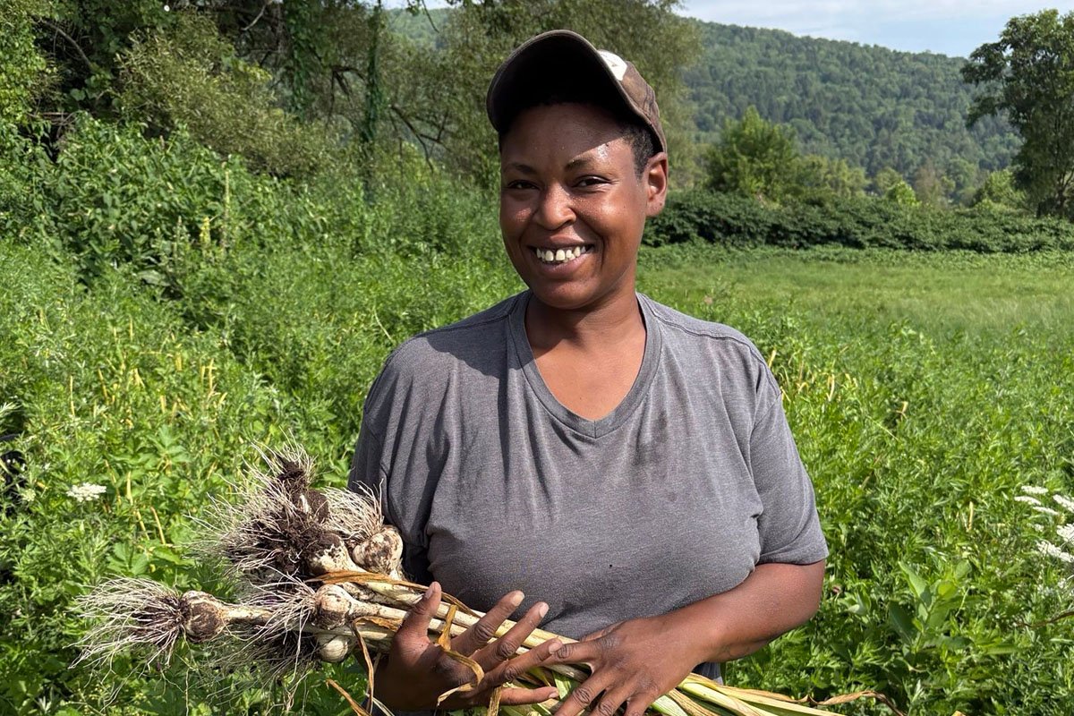 A Black woman with cropped hair, a billed cap, and a welcoming smile stands in a verdant field holding a harvest of garlic.