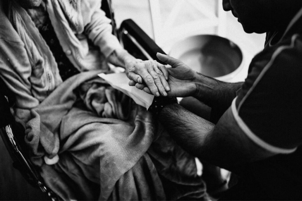 A caregiver holds an elderly person's hand in their lap, photographed in black and white, a wheelchair visible in the background.