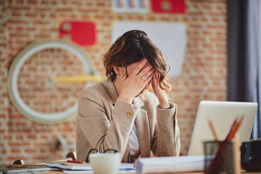 A woman with a curled bob sits at her work desk and buries her head in her hands, in exhaustion and frustration.