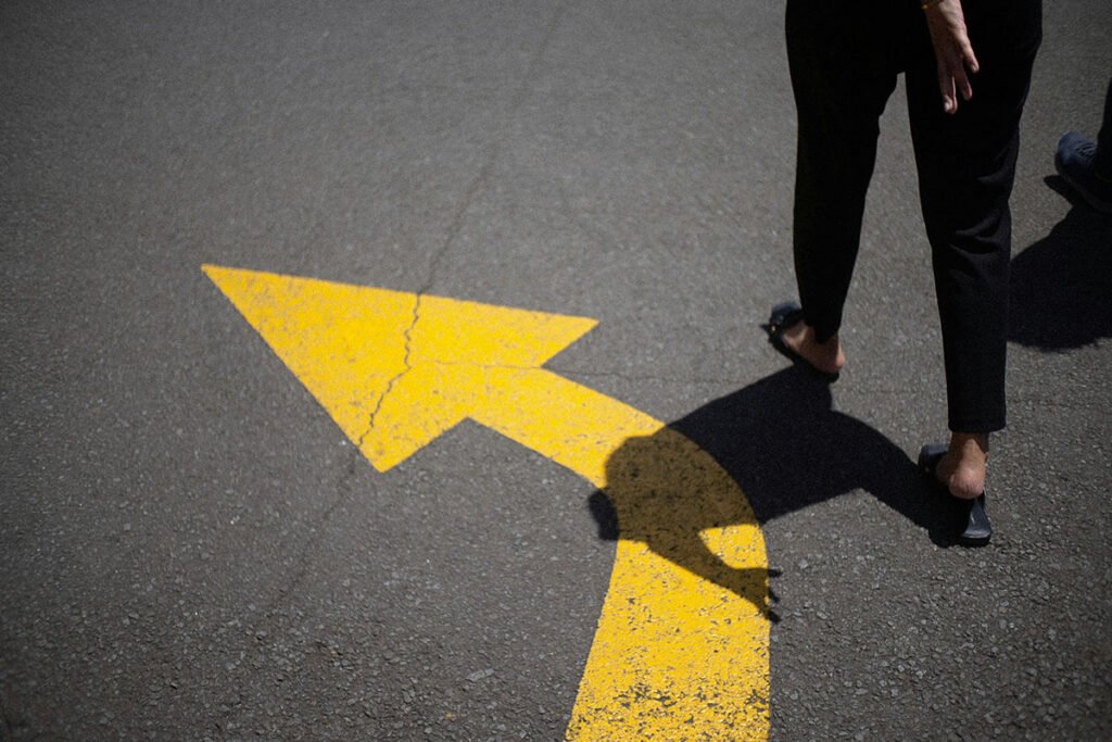 A cropped photo of a person’s legs walking along a paved road. There is a yellow arrow veering to the the side and pointing ahead. The person walks alongside this arrow.