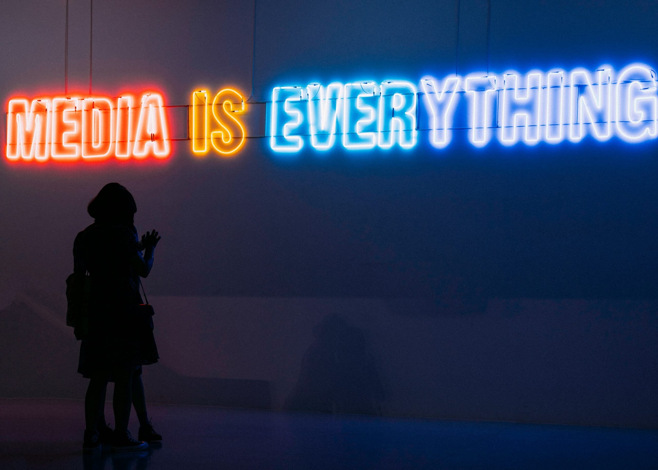 Silhouette of two people standing in front of a neon sign that reads, "Media Is Everything."