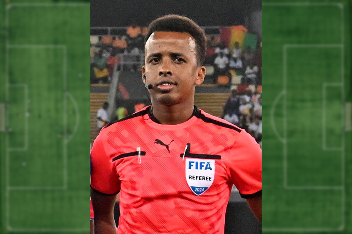 Referee Omar Abdulkadir Artan wearing a red referee shirt with stands of fans behind him. Imposed on an image of a soccer field.