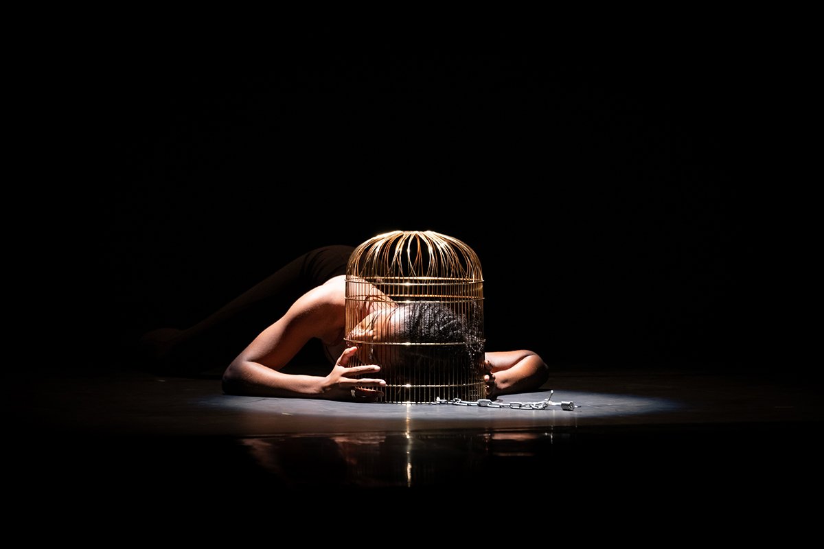 A Black person lies on the floor of a stage with a birdcage around their head. The cage has a chain attached and a spotlight lights the scene.