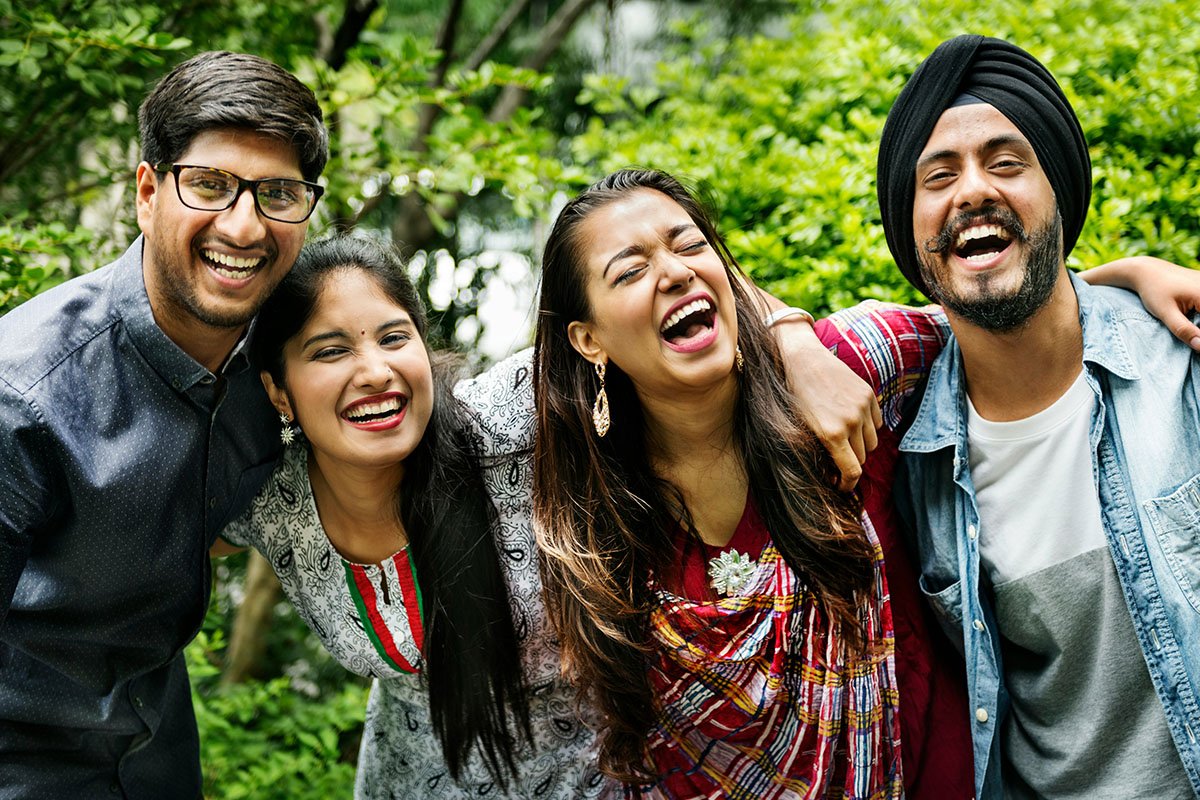 A group of four southeast Asian people with arms around each other, laughing together. The man on the end wears the Sikh Dastar (pagri).