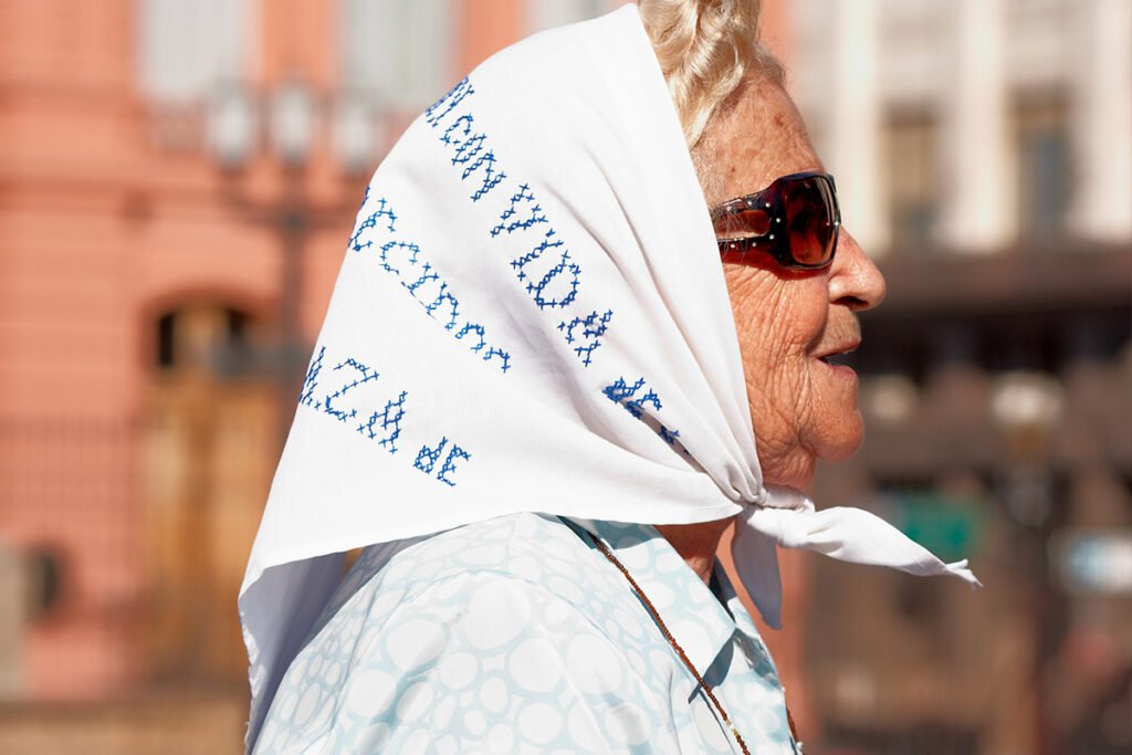 Mercedes Colás de Meroño, an elderly woman in profile wearing sunglasses and the signature white shawl of the Mothers of Plaza de Mayo.