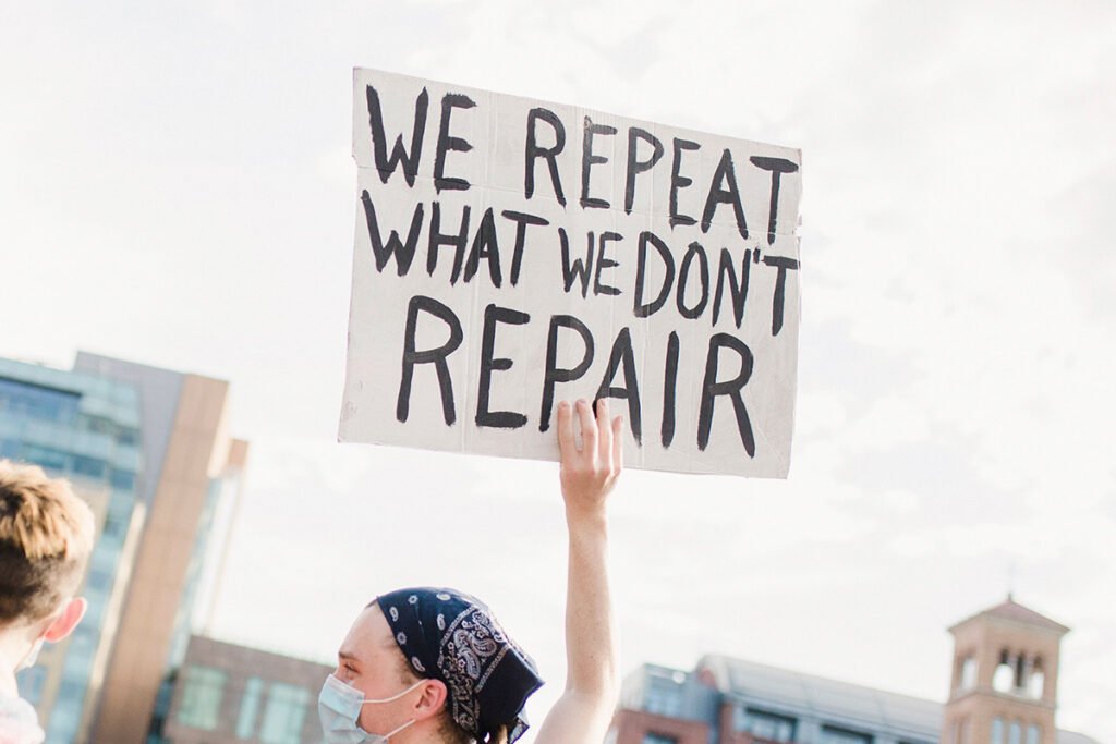 A person holds up a hand-painted protest sign that reads, “We repeat what we don’t repair”
