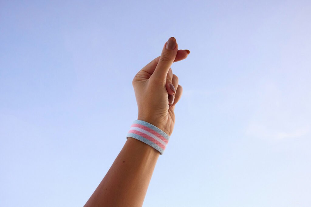 A person with mauve colored nails and a trans flag wristband holding their hand up in a songarak-hateu (korean finger heart).