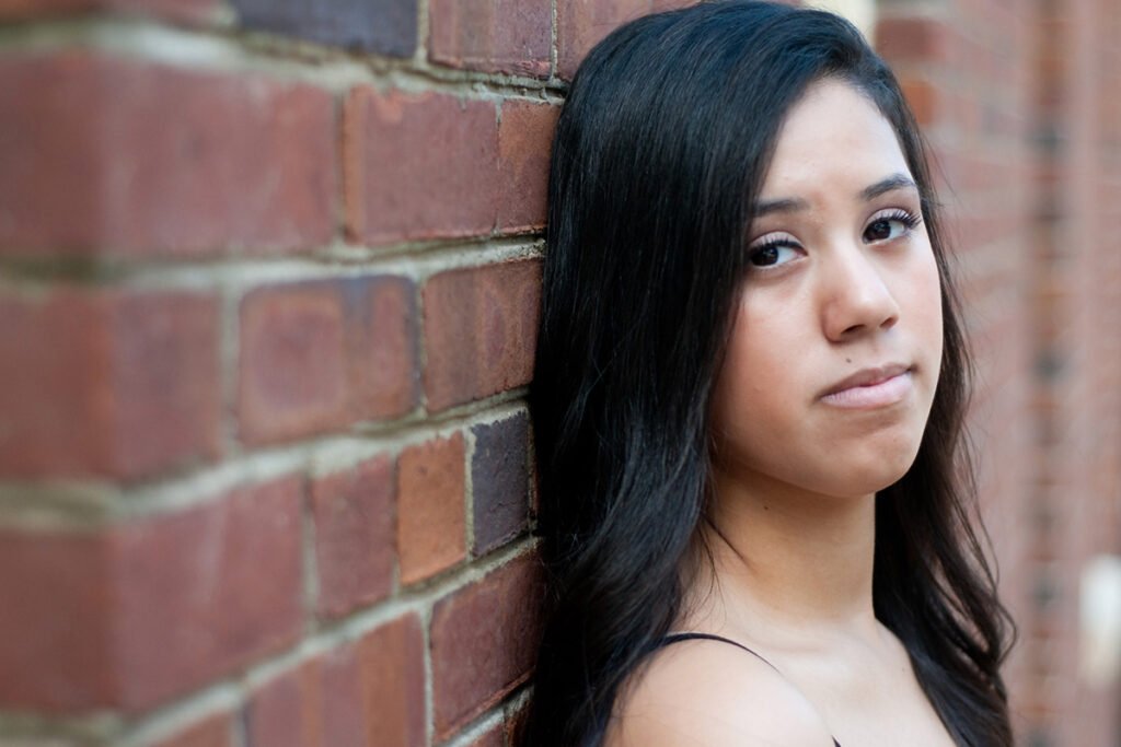 A teenage girl with brown skin leans up against a brick wall and looks into the camera with a serious face.