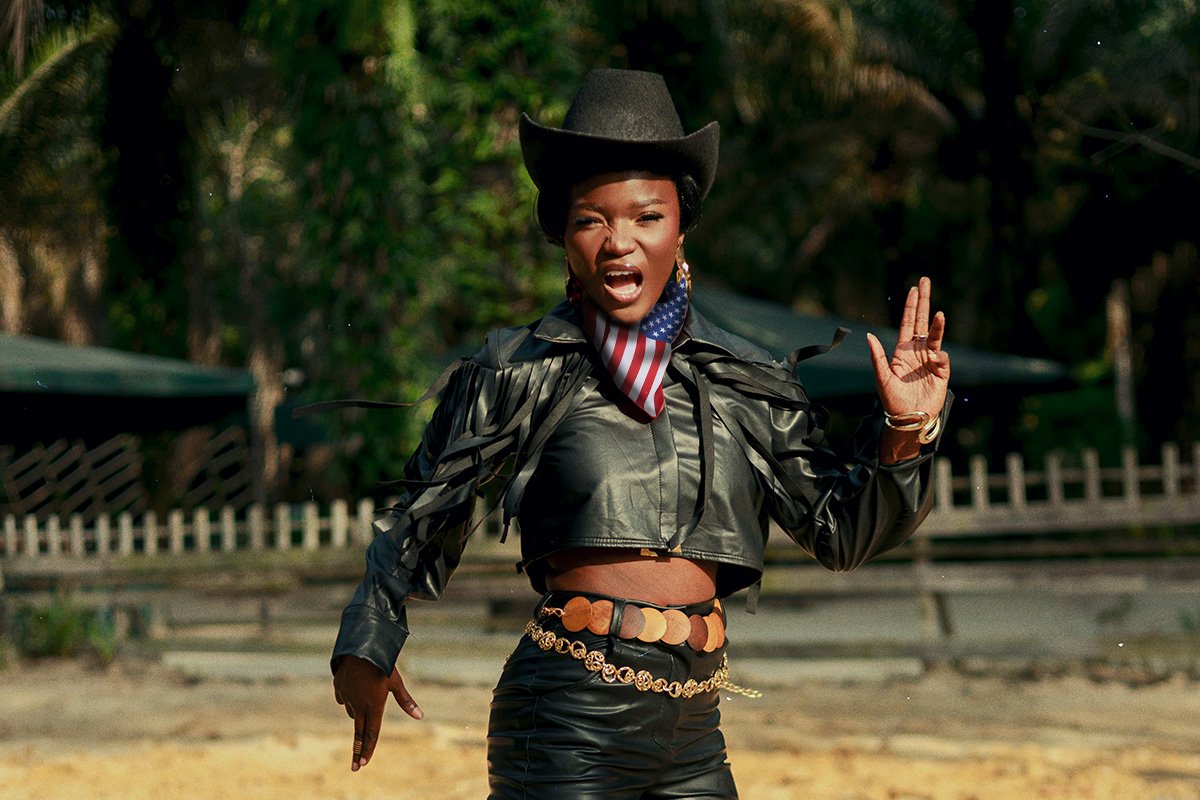 A Black woman with short hair wears a typical cowgirl outfit featuring a Black leather hat and cropped jacket, with a decorative belt and an American flag bandana around her neck.