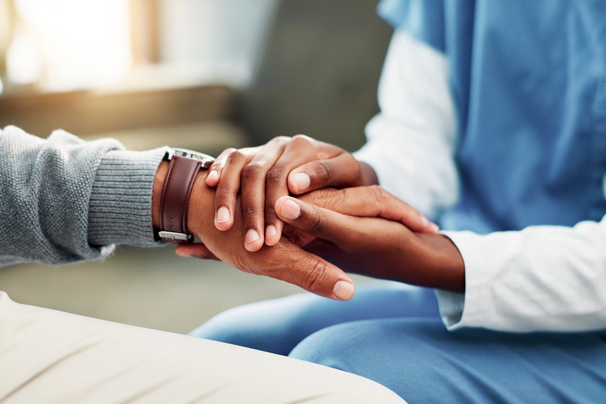 A close-up of two pairs of hands holding onto each other, one pair of hands belongs to a Black woman wearing a stethescope and blue scrubs. The other hand belongs to a Black man with a watch.