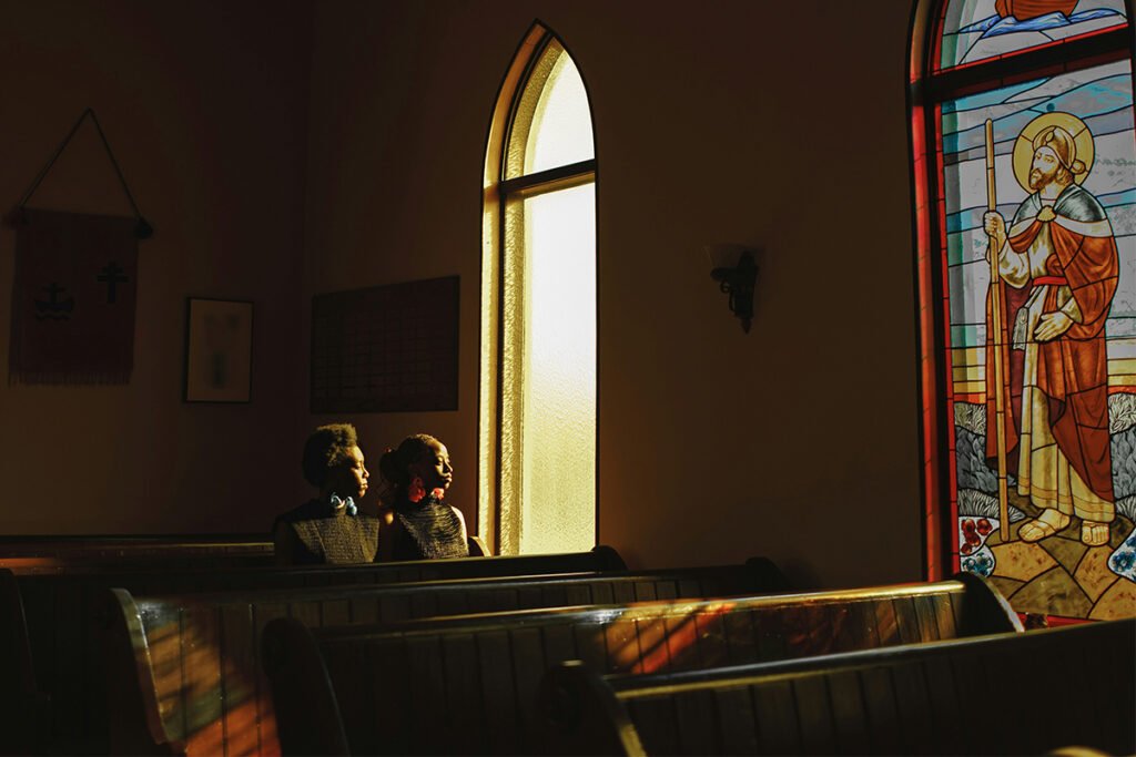 Two Black women sit in the pews of a dimly lit church. They look to the left, where there is a stained glass window showing a religious figure.