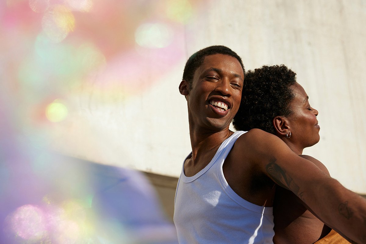 A Black man and woman dance together, back-to-back with arms outstretched and with smiles on their faces. In the foreground, an iridescent fabric covers the lens, giving it a magical, colorful effect.