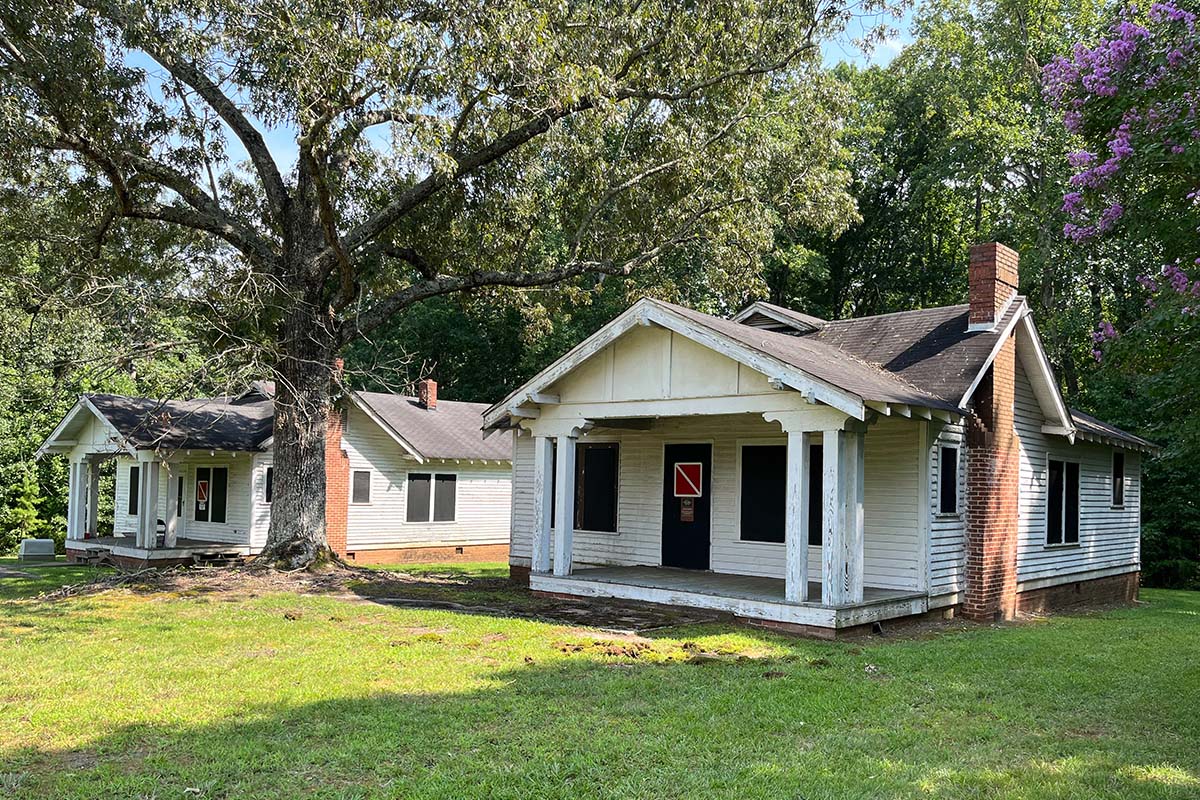 Two worn cottage-style buildings with white wooden exteriors and brick chimneys. They stand side-by-side in a wooded area.