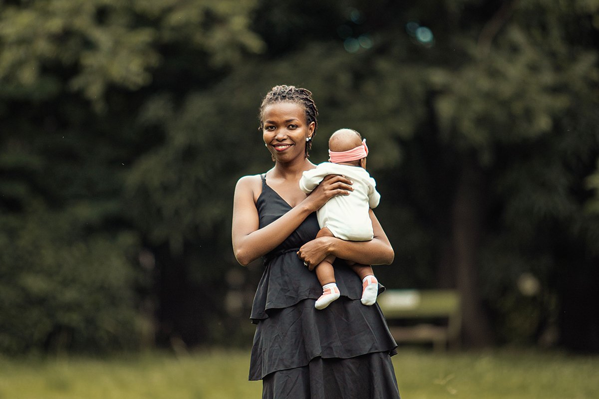 A Black woman with a black dress stands in a field holding a baby girl in a onesie and bow headband. The mother smiles softly.