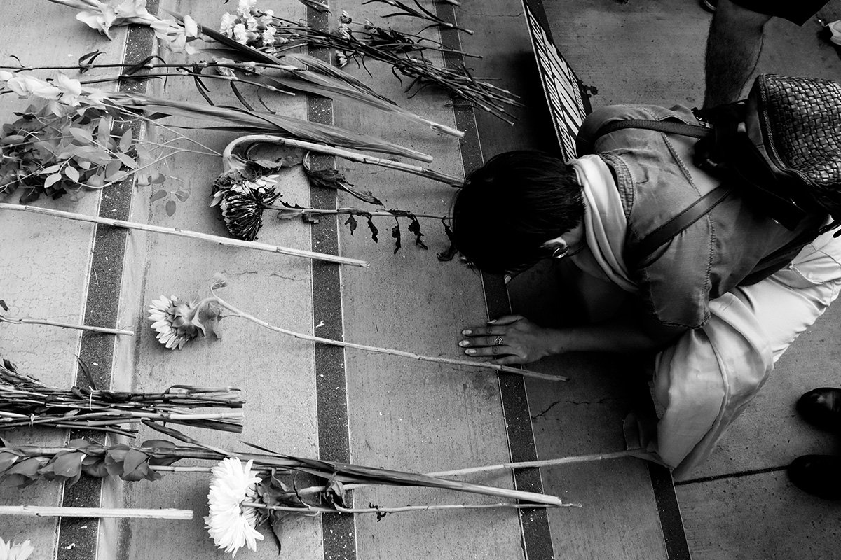 A black and white photo of a man on his knees praying on concrete steps. Before him are many flowers laid upon the steps.