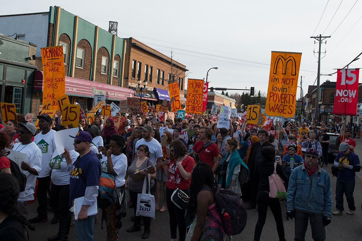 A photo showing a Strike and a protest, with a crowd of people on a street holding signs demanding a $15 minimum wage. Dinkytown, Minnesota.